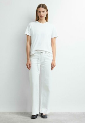 Woman standing against white wall wearing a plain white t-shirt, white drawstring pants, and black square-toe shoes.