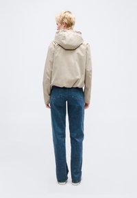 Άτομο με κοντά ξανθά μαλλιά, που φοράει μπεζ μπουφάν με κουκούλα και μπλε τζιν, στέκεται γυρισμένο με την πλάτη προς τα εμπρός σε απλό λευκό φόντο.