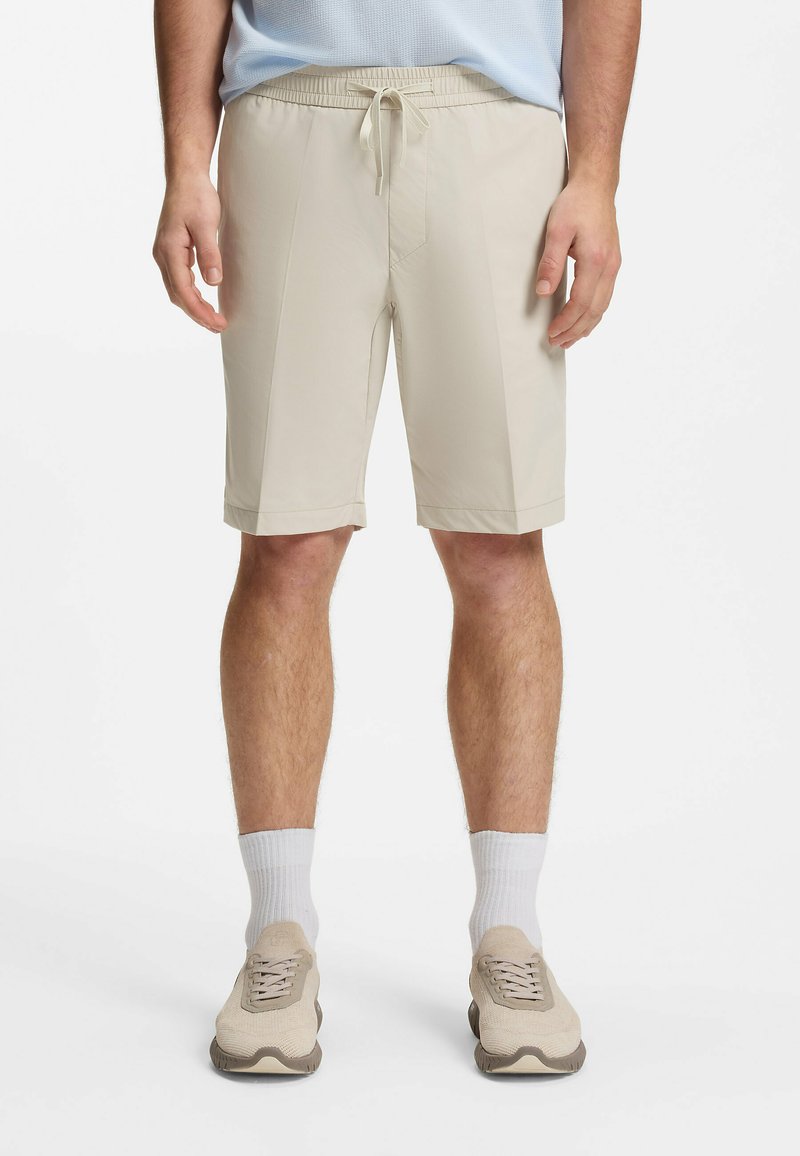 Homme portant un short beige mi-long avec taille à cordon, chaussettes blanches et baskets beiges sur un fond uni.