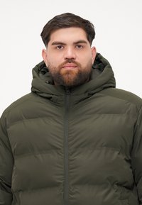 Man with short dark hair and beard wearing a zipped dark green padded jacket with hood against plain background.