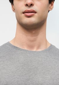 Κοντινή λήψη του κάτω μέρους του προσώπου και του λαιμού ενός νεαρού άνδρα που φοράει γκρι μπλουζάκι με στρογγυλή λαιμόκοψη, πάνω σε απλό φωτεινό φόντο.