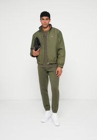 Καφέ μπόμπορ τζάκετ με φερμουάρ μπροστά και ελαστικούς καρπούς, συνδυασμένο με αντίστοιχους φόρμες. Το μοντέλο κρατά μια μαύρη τσάντα και φοράει λευκά αθλητικά παπούτσια.