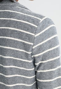 Grå och vit randig tyg, texturerad stickning, med krage och avslappnad passform. Detaljer inkluderar växlande randbredder.