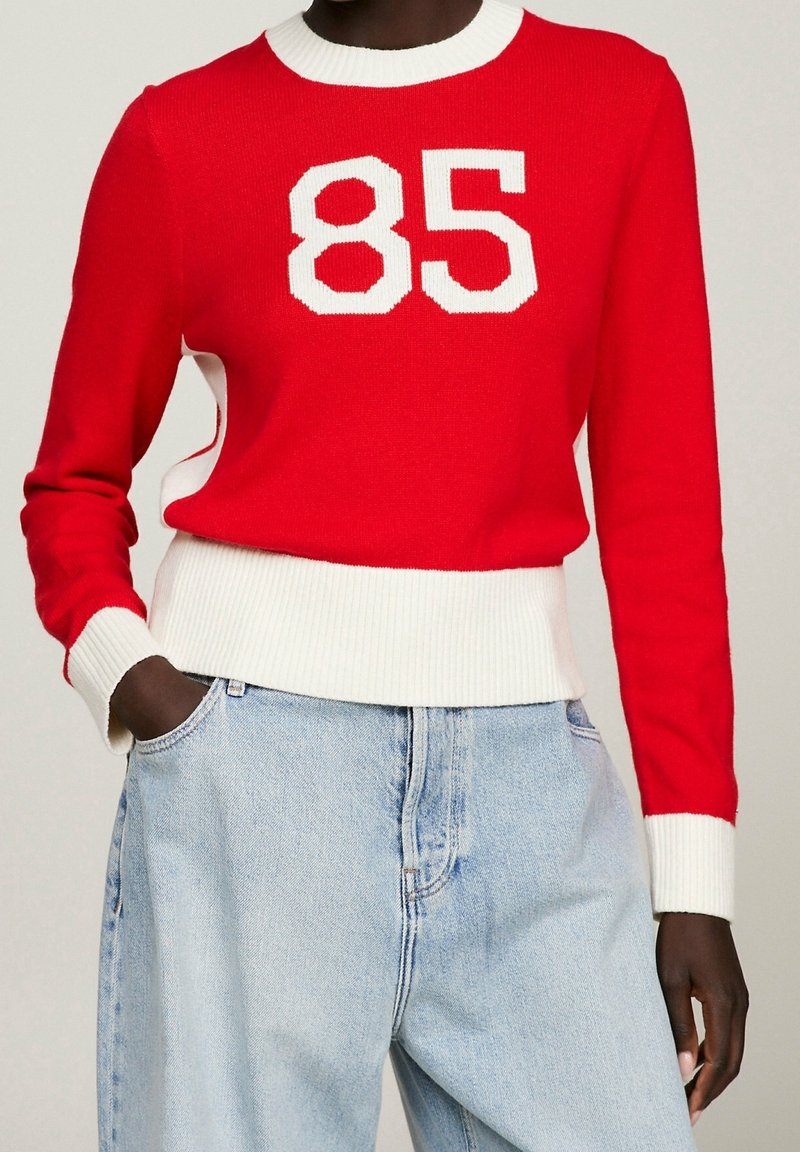 Pull rouge à manches longues avec col, poignets et ceinture côtelés blancs, arborant le numéro blanc "85" sur la poitrine, porté avec un jean bleu clair.