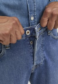 Jeans de denim azul con cierre de botón, que cuentan con herrajes metálicos, costuras amarillas y un primer plano de manos abrochando la cintura.