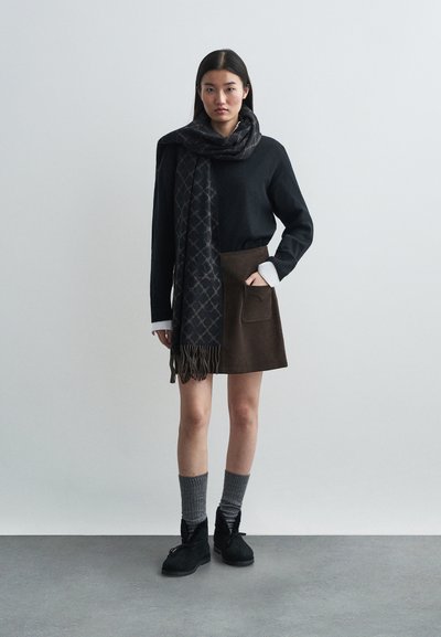 Pull noir, jupe marron avec poche, écharpe sombre à motifs avec franges, chaussettes grises, chaussures noires. Design simple et moderne avec des textures variées.