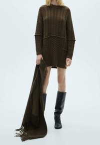 Robe pull surdimensionnée en vert olive foncé avec des motifs texturés, assortie à un châle frangé et des bottes noires montantes.