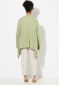 Grön oversized cardigan i bladgrön färg med hög-låg nederkant, stickad textur, öppen framsida, bärs över beige byxor, kombinerad med nude höga klackar.