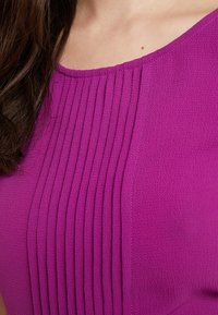 Detailní záběr na osobu v purpurovém strukturovaném topu s vertikálními sklady, vidět část výstřihu a dlouhé hnědé vlasy.