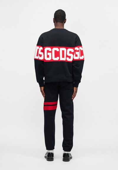 Μαύρη μπλούζα με μεγάλο �λευκό και κόκκινο λογότυπο, συνδυασμένη με μαύρο παντελόνι που έχει κόκκινες ρίγες στο μηρό. Το μοντέλο στέκεται γυρισμένο προς τα πίσω.