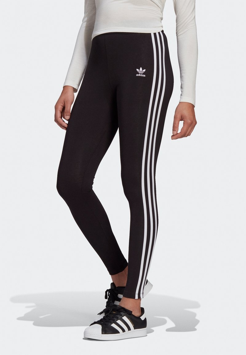 Osoba ubrana w czarne legginsy Adidas z białymi trzema paskami po bokach oraz czarne sneakersy Adidas z białymi paskami.