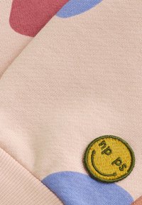 Dzianina w miękkim różowym kolorze z niebieskimi i czerwonymi zaokrąglonymi kształtami. Posiada okrągłą żółtą naszywkę z haftowanym uśmiechniętym emotikonem i tekstem.