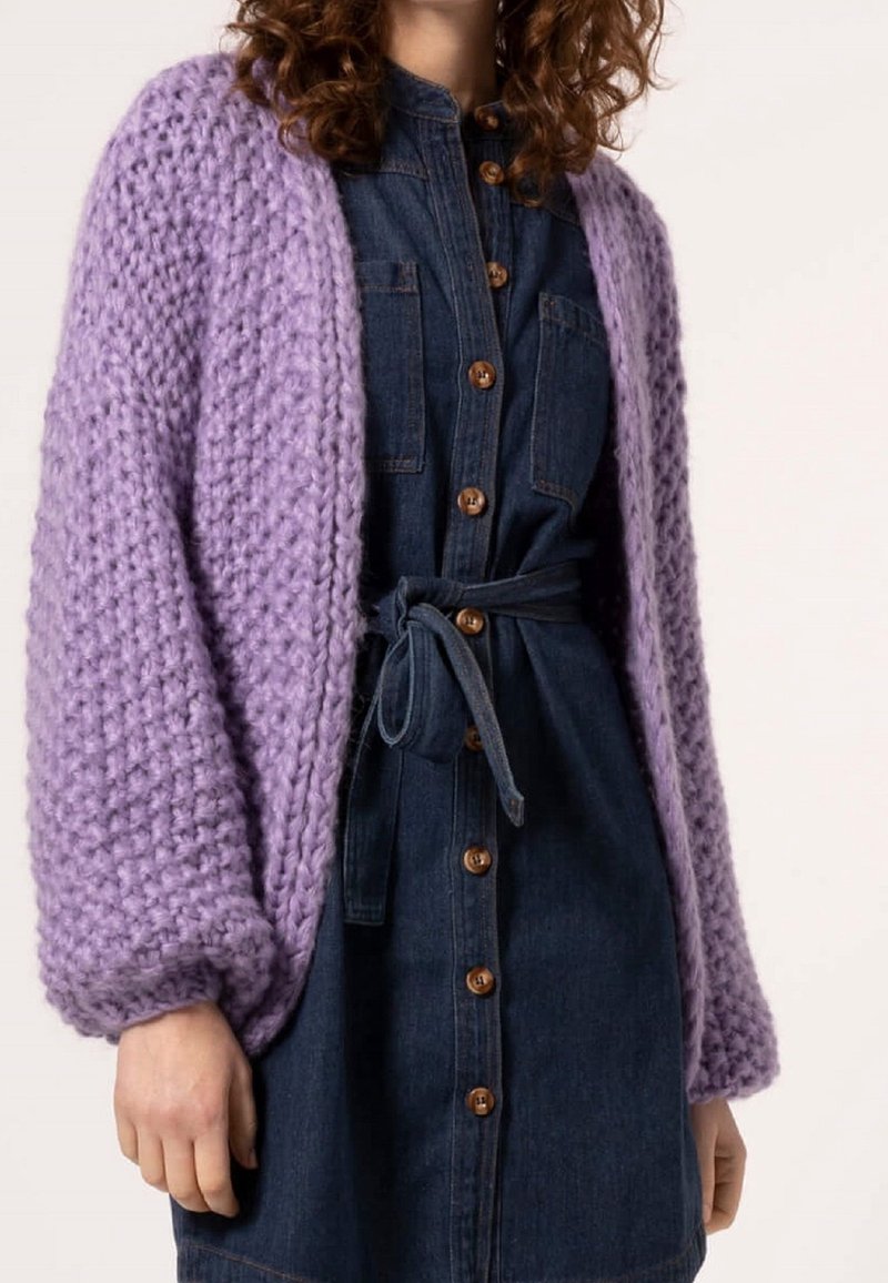 Femme portant un gros cardigan tricoté lilas sur une robe en denim foncé boutonnée avec ceinture, vue des épaules jusqu'à mi-cuisse.