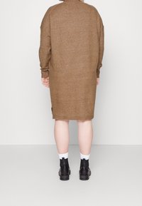 Robe marron en tricot avec col haut, manches longues, fentes latérales et poignets côtelés. Portée avec des bottines noires et des chaussettes blanches.