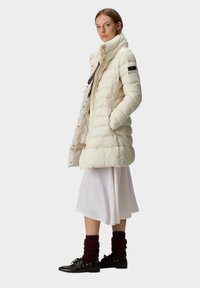 Gewatteerde witte donsjas met een hoge kraag en ritssluiting, voorzien van een logopatch op de mouw en goudkleurige details. Draagt een witte jurk en bordeauxrode beenwarmers.