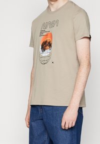 Beige bomulds-t-shirt med korte ærmer med et NASA-grafik, tekst og et orange og sort designelement, kombineret med blå jeans.
