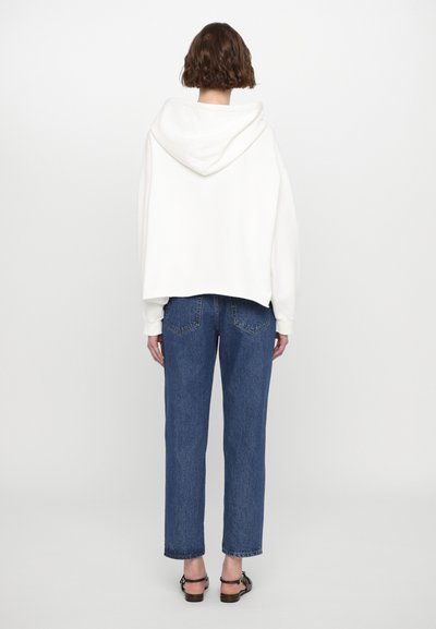 Λευκό hoodie με κα cropped σχεδίαση, σε συνδυασμό με τζιν παντελόνι ψηλής μέσης σε μπλε απόχρωση. Το υλικό φαίνεται μαλακό, με λεία υφή.