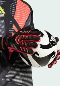 Black and red goalkeeper gloves with textured grip hold a black and white soccer ball. The gloves feature a striking design with finger protection accents.