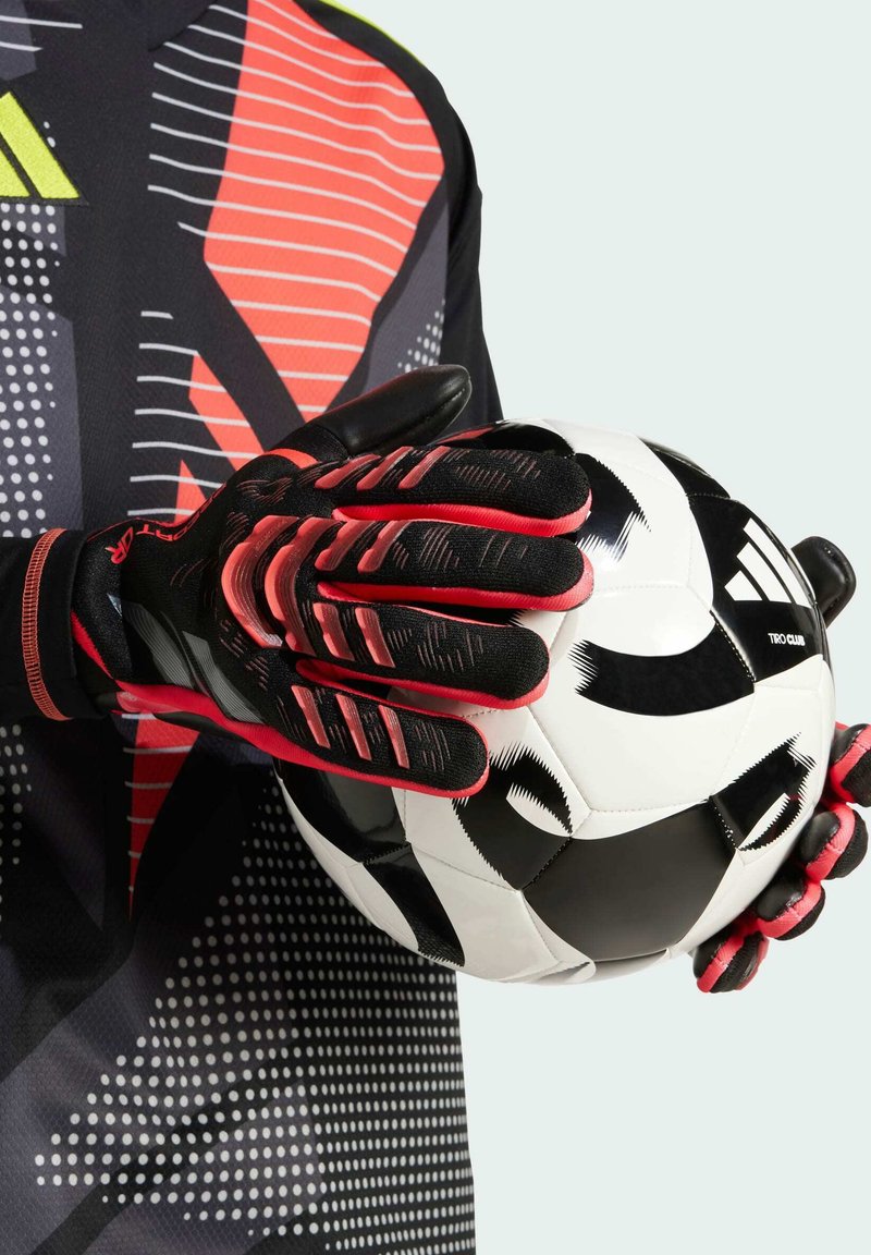 Schwarze und rote Torwarthandschuhe mit strukturiertem Griff halten einen schwarz-weißen Fußball. Die Handschuhe weisen ein auffälliges Design mit Fingerschutzakzenten auf.