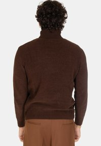 Maglione a maglia marrone con colletto alto, polsini e orlo a coste, caratterizzato da una texture liscia. Indossato con pantaloni marroni chiari, visto da dietro.
