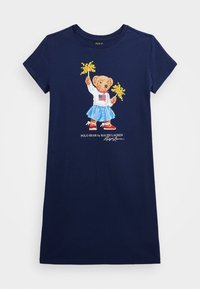 Marinblå T-shirt med en björn i en vit topp med en amerikansk flagga och en blå kjol, som håller gula pom-poms. Mjuk bomullstextil.
