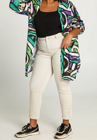 Chemise oversize colorée avec des motifs abstraits en vert, violet et noir. Associée à un pantalon ajusté beige clair et des baskets noires.