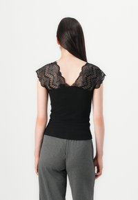 Canotta nera in cotone con maniche a cappuccio in pizzo e design a V sul retro. Abbinata a pantaloni grigi, caratterizzati da una texture morbida.