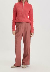 Korallinvärinen, teksturoitu neulepaita, jossa on puolivetoketjukaulus, yhdistetty korkeavyötäröisiin, löysälahkeisiin vaaleanpunaisiin housuihin. Beigejä kenkiä täydentämään asua.