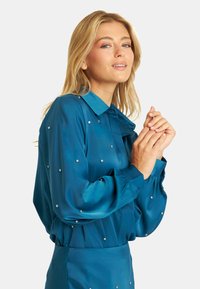 Blusa verde azulado con una textura suave, que presenta un collar, mangas abullonadas y acentos de strass dispersos en un diseño ajustado.