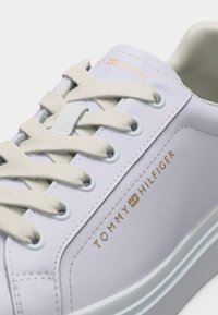 Baskets en cuir blanc avec lacets beige et logo Tommy Hilfiger doré sur le côté, présentées en gros plan.