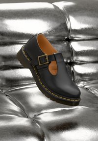 Chaussure en cuir noir avec un design découpé et une boucle dorée. Elle présente une semelle marron texturée et une couture jaune le long du bord.