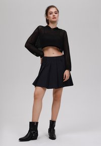Maglione corto nero a maglia con cappuccio, abbinato a una gonna plissettata nera a righe. Il modello indossa stivaletti neri alla caviglia con accentuazioni in argento.