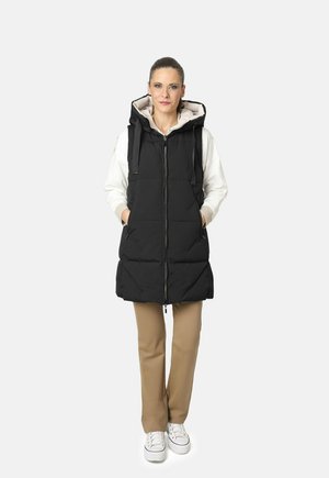 Chaleco de plumas negro con capucha, que cuenta con cierre en la parte delantera, bolsillos laterales y un diseño acolchado, combinado con pantalones beiges y zapatillas blancas.