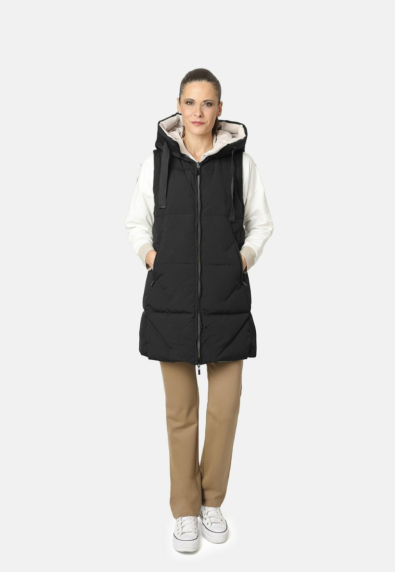 Chaleco de plumas negro con capucha, que cuenta con cierre en la parte delantera, bolsillos laterales y un diseño acolchado, combinado con pantalones beiges y zapatillas blancas.