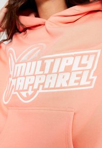 Korálová mikina vyrobená z měkké tkaniny s přední kapsou, na které se nachází výrazné bílé logo "MULTIPLY APPAREL" a sportovní grafické detaily ve tvaru motýla.