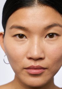 Nahaufnahme eines Gesichts mit glatter, heller Haut, dunklen braunen Augen, natürlichem Make-up und einem silbernen Creolenohrring. Einfacher Hintergrund.