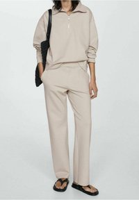 Felpa beige con zip e colletto, abbinata a pantaloni larghi coordinati. Realizzata in tessuto morbido, dotata di tasche laterali. Borsa nera intrecciata.