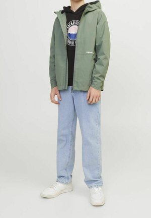 Person iført en lysegrøn hættejakke over en sort grafisk hoodie, lyseblå jeans og hvide sneakers, stående mod en ensfarvet baggrund.