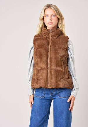 Blonde vrouw die een bruin geribd vest draagt over een gestreept shirt met lange mouwen en een blauwe spijkerbroek, staand tegen een effen achtergrond.