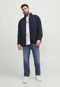 Marineblå pufferjakke med quiltet front, lange ærmer og høj krave. Bæres over en hvid grafisk t-shirt og parret med blå jeans og hvide sneakers.