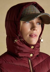 Punane polsterdatud jakke kõrge kraega ja kuldsete detailidega, täiendatud mustrilise mütsiga maapinnast inspireeritud toonides. Tekstuuriga kangas ja quiltitud disain on nähtavad.