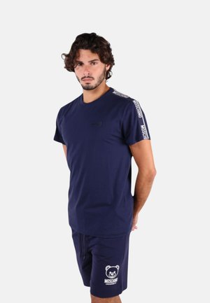 Uomo con capelli ricci che indossa una t-shirt Moschino blu navy e pantaloncini abbinati con logo orsetto, in piedi su sfondo bianco.