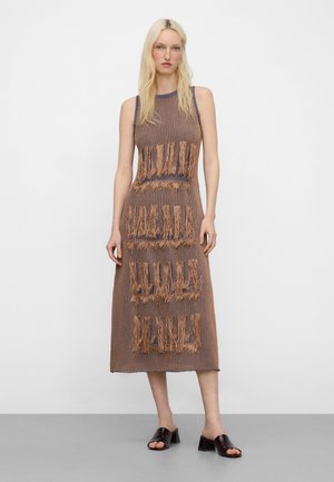 Donna bionda che indossa un vestito marrone senza maniche a maglia con pannelli orizzontali sfrangiati e sandali neri con tacco largo e punta aperta, in piedi contro un muro bianco.