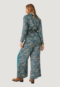 Mameluco de estampado floral en verde azulado con mangas largas, cintura ceñida, pantalones anchos y acentos en los paneles laterales. Botines marrones completan el look.