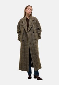 Manteau en laine à carreaux marron avec un devant double boutonnage, des revers larges et des poches latérales, associé à un jean bleu et des chaussures sombres et brillantes.