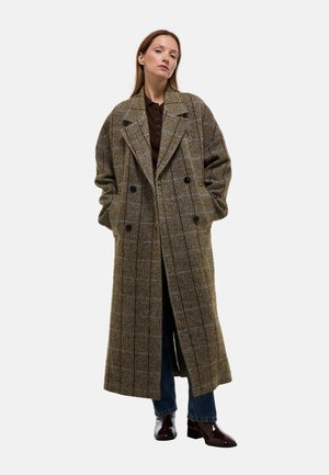 Manteau en laine à carreaux marron avec un devant double boutonnage, des revers larges et des poches latérales, associé à un jean bleu et des chaussures sombres et brillantes.
