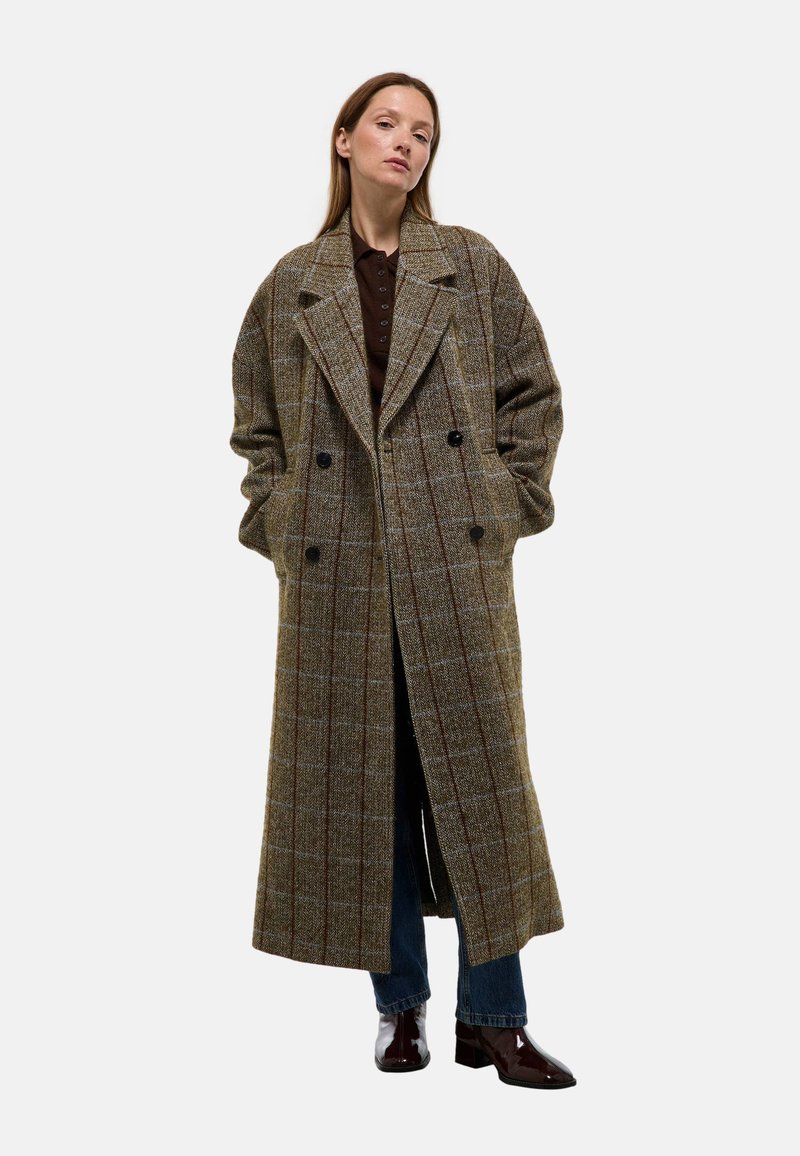 Manteau en laine à carreaux marron avec un devant double boutonnage, des revers larges et des poches latérales, associé à un jean bleu et des chaussures sombres et brillantes.