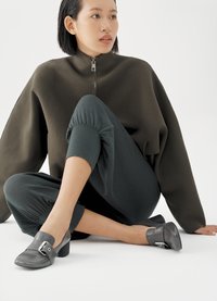 Graue Lederschuhe mit abgerundetem Zehenbereich, ausgestattet mit einem verstellbaren Riemen und silbernen Beschlägen, kombiniert mit einem dunklen Pullover und passenden Hosen.