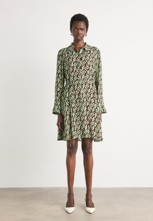 Groene en bruine jurk met een patroon, voorzien van een kraag en lange mouwen, met ruffles aan de manchetten en een uitlopende silhouet.