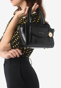 Borsa nera lucida con doppie maniglie, tasca anteriore, dettagli dorati e logo del marchio. Design compatto con angoli arrotondati e cinque piedi in metallo.
